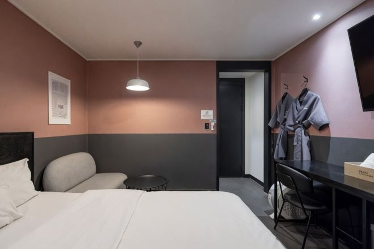 A hotel room with a bed, a desk, a chair, a robe rack, and a door.