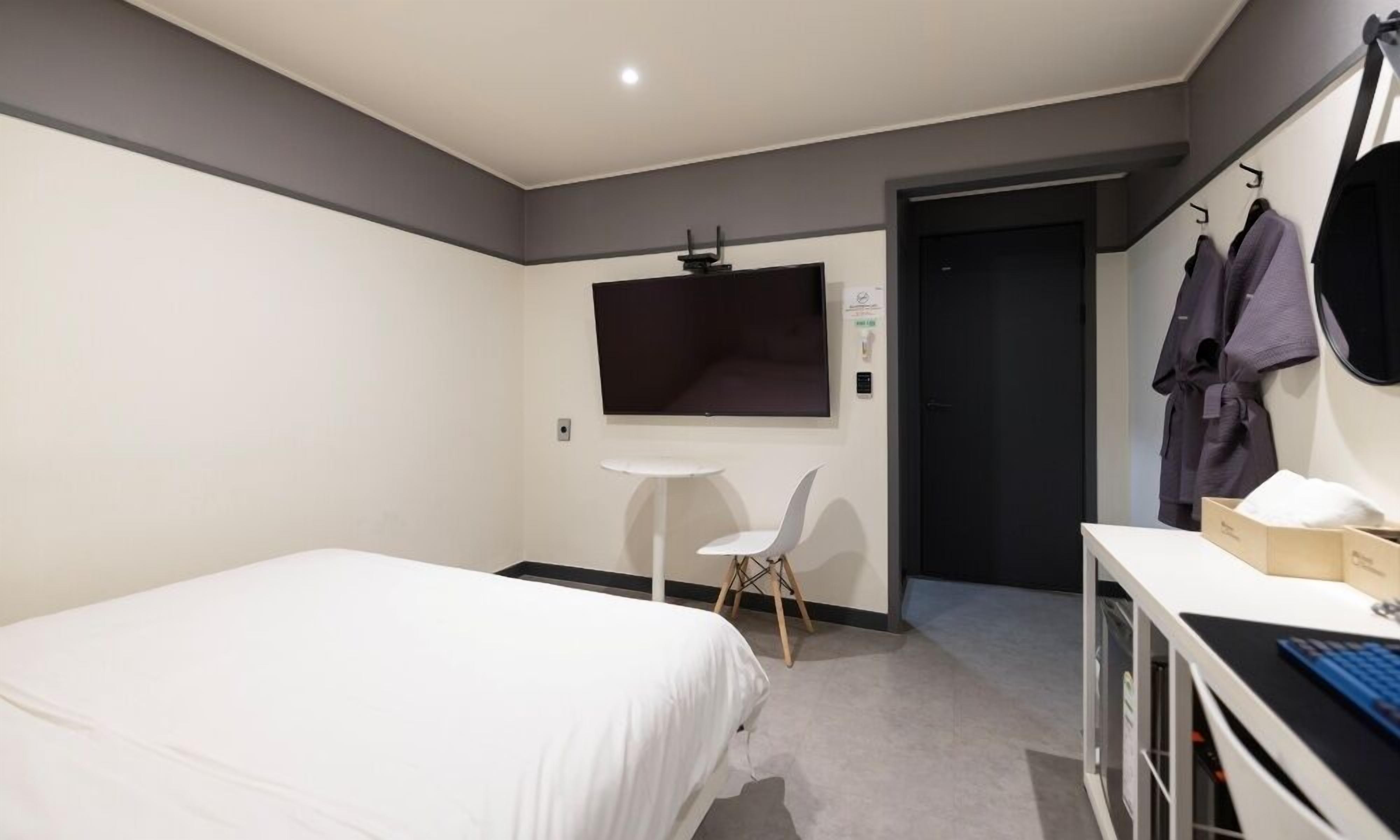 A hotel room with a bed, a small desk with a computer, a chair, a TV mounted on the wall, and a door leading to another room.