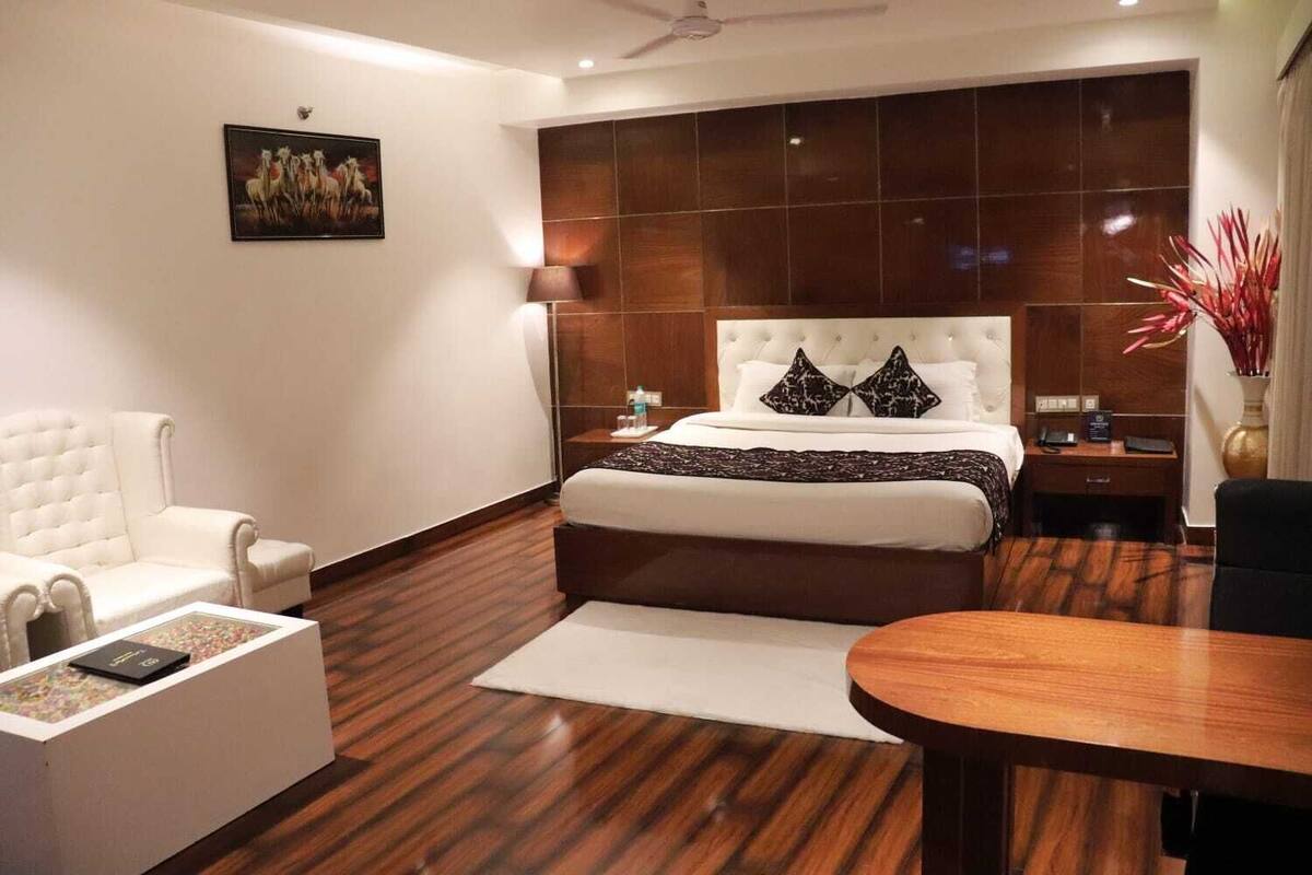 A modern hotel room with a large bed, a white armchair, a wooden coffee table, and a desk.