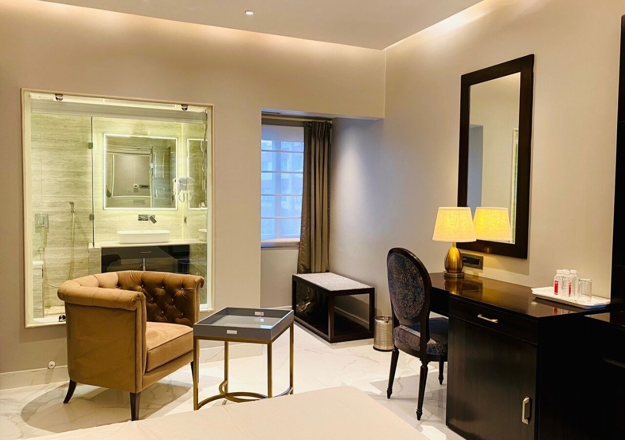 A hotel room with a sofa, a small table, a chair, a desk, a mirror, and a bathroom with a glass shower enclosure.