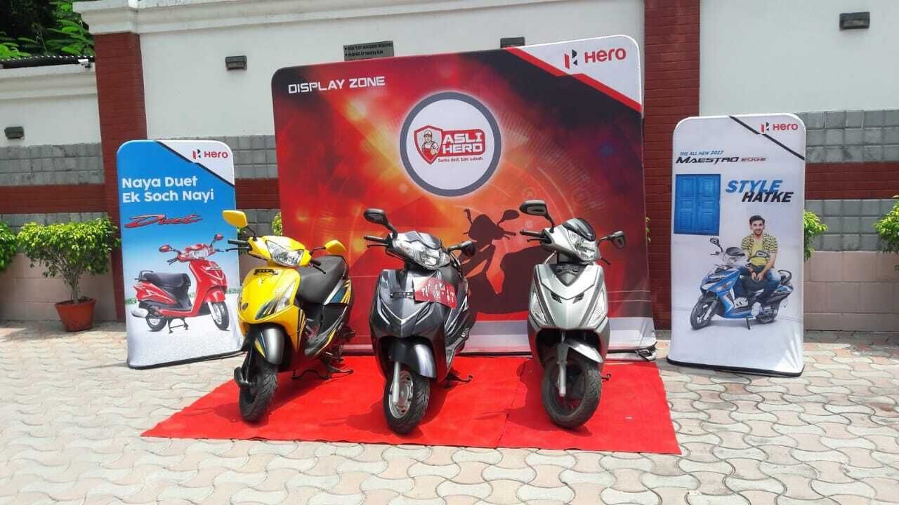 Tres motocicletas estacionadas frente a un espacio de exhibición con pancartas que anuncian 'Hero' y 'ASLI HERO'.
