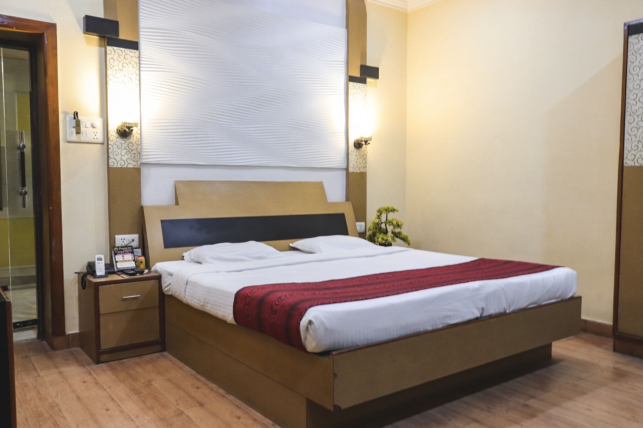 A neatly made bed with a red blanket, a bedside table with a phone and a lamp, and a small plant on the nightstand.