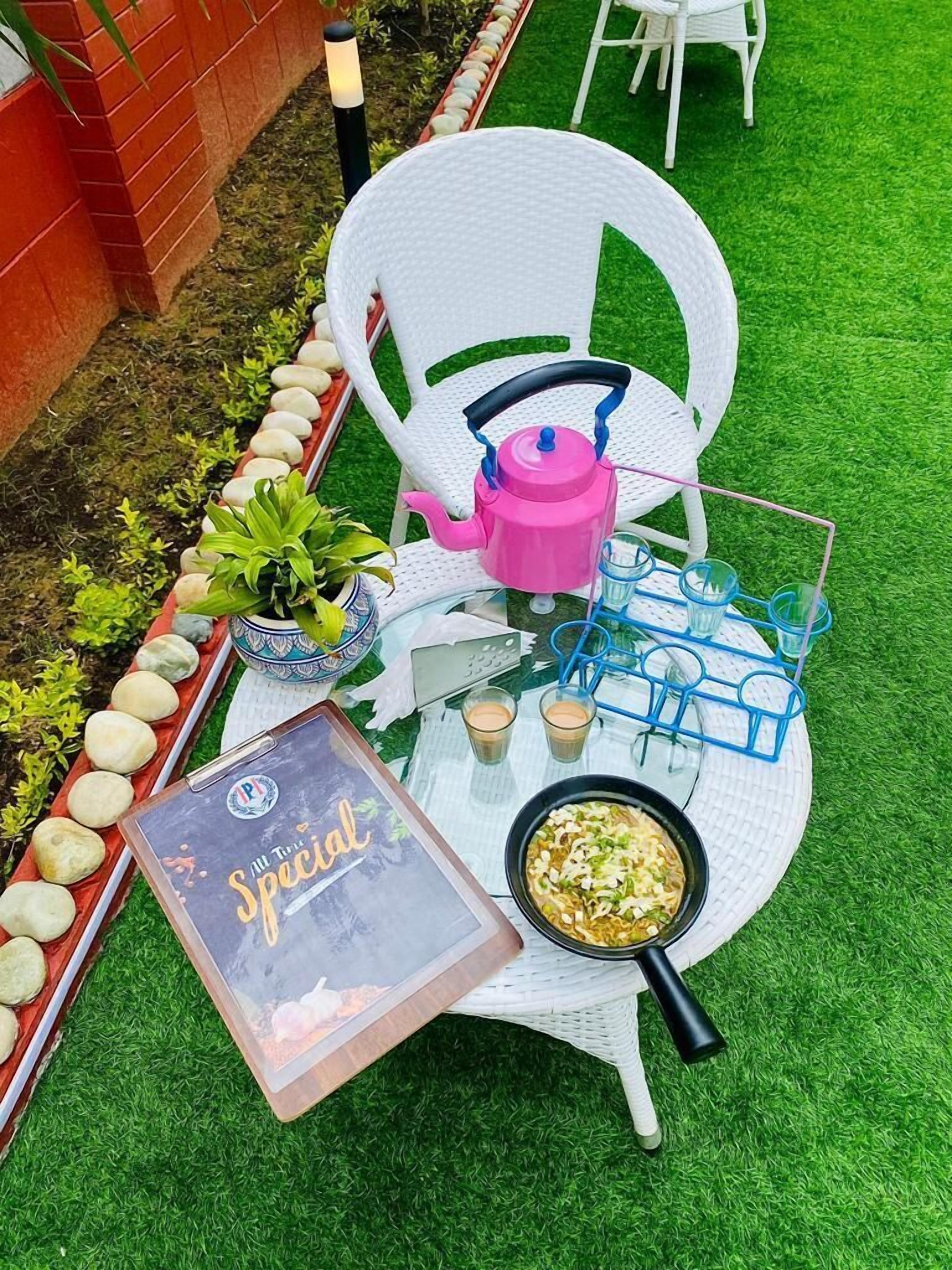 A white outdoor table with a pink kettle, two glasses, a bowl of food, a book titled 'All the Special', and a potted plant.