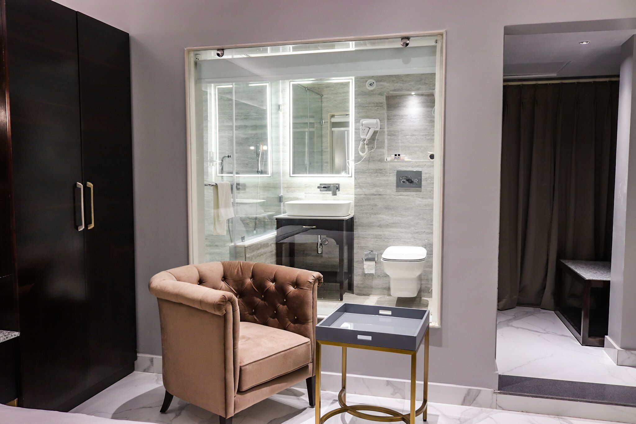 Une salle de bain moderne avec une douche à parois de verre, un meuble-lavabo et des toilettes. On y trouve également un fauteuil à capitons et une petite table avec un plateau en verre.
