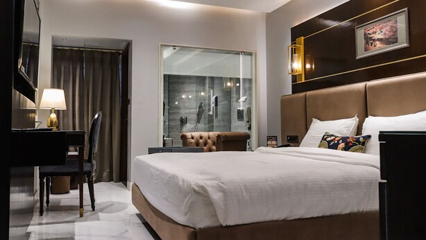 A modern hotel room with a large bed, a desk with a lamp, a chair, and a glass shower enclosure. - Prayag Inn (Prayagraj)