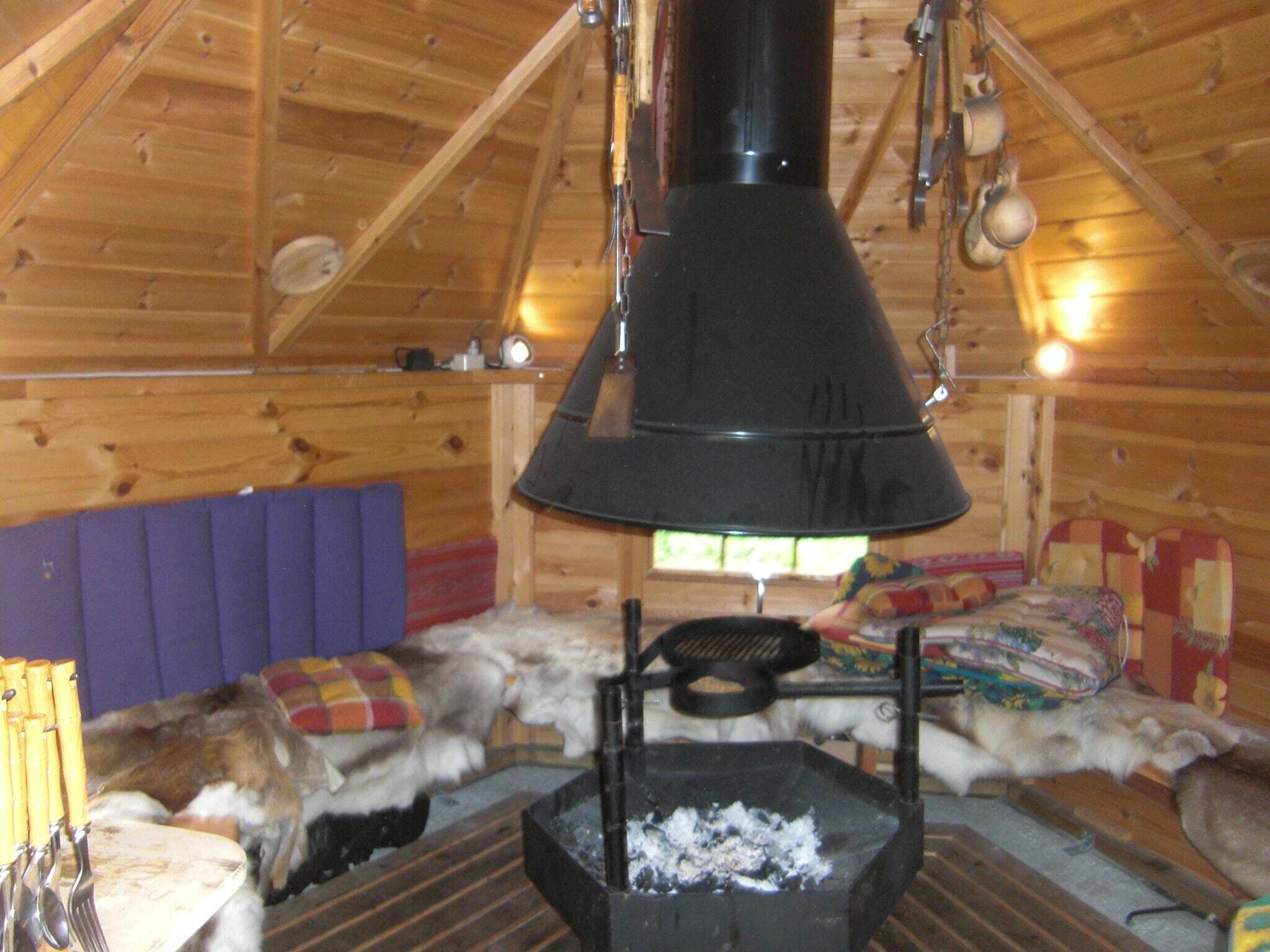 A cozy cabin interior with a wood stove, a purple cushioned bench, and a sleeping area with a colorful blanket.