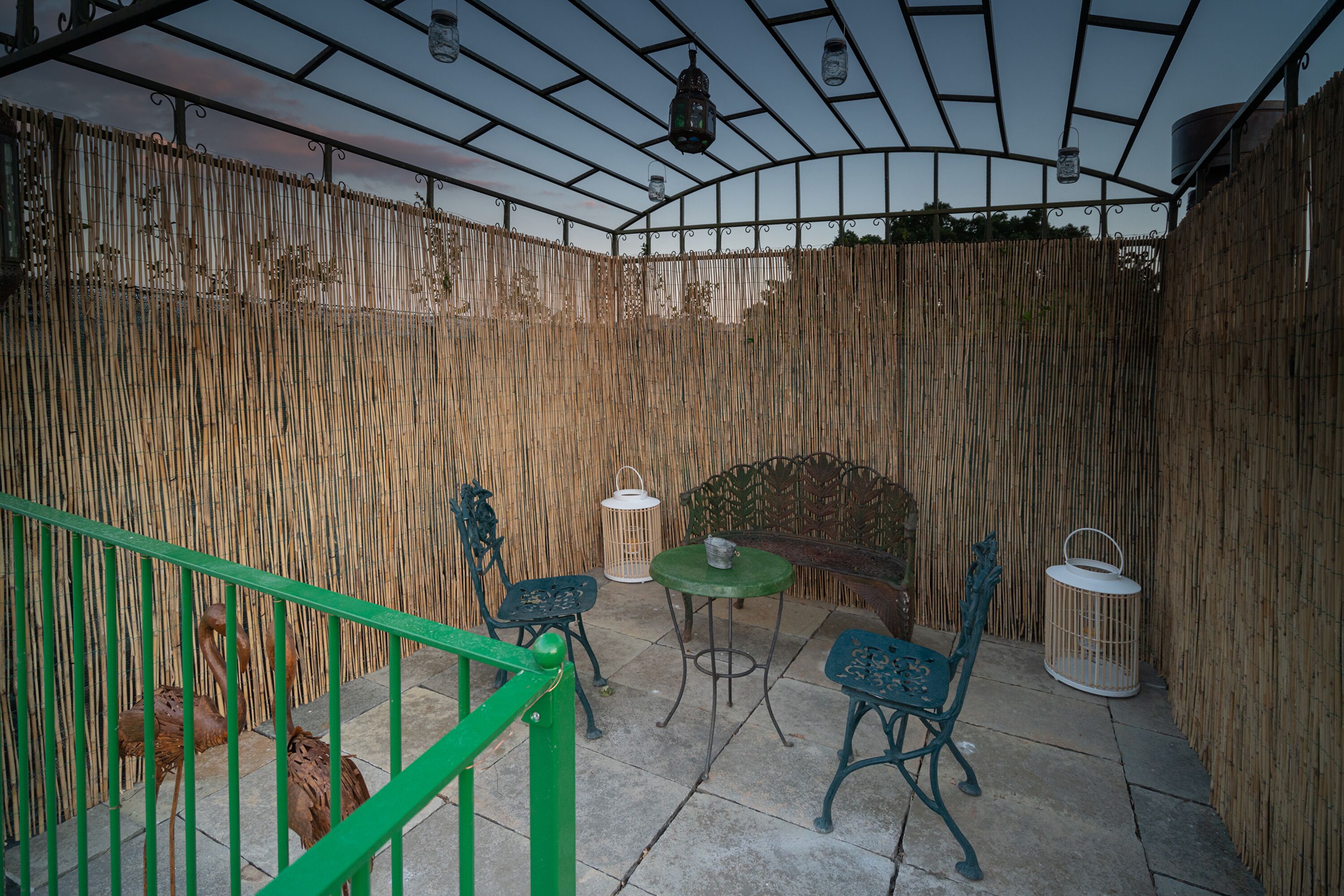 A covered outdoor patio with bamboo walls, green metal chairs, a small table, and hanging lanterns.