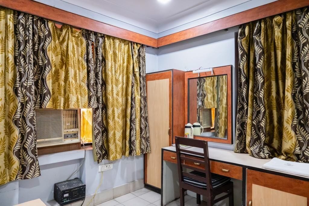 A room with a desk, chair, and a mirror. There is a window with curtains, a radiator, and a small shelf with items.