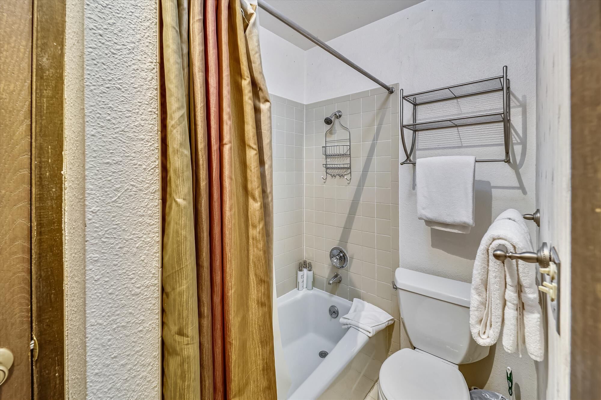 Et badeværelse med badekar, toilet og bruser. Der er håndklæder hængende på væggen og en hylde med toiletartikler.