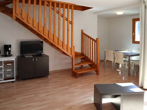 A modern living room with a wooden staircase, a flat-screen TV, a coffee maker, and a dining area with chairs and a table.