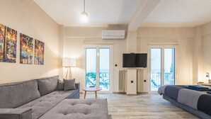 En moderne stue med en grå sofa, et lille rundt bord og et TV ophængt på væggen. To skydedøre i glas fører ud til en balkon med udsigt.
