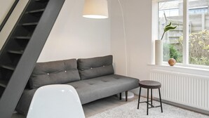 A modern living room with a grey sofa, a white chair, a small round stool, a hanging lamp, and a window with a view of greenery. - Beach front home in Bergen aan Zee (Bergen aan Zee)