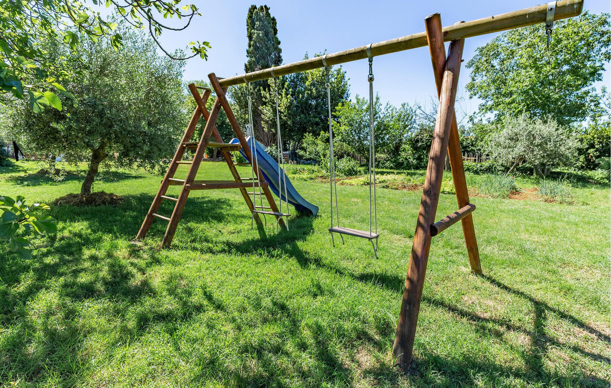 A wooden swing set in a grassy area with trees and a slide in the background.
