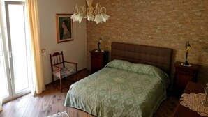 Une chambre avec une couvre-lit à motifs verts, une chaise en bois, une table de chevet en bois avec une lampe, un lustre et un mur en briques.