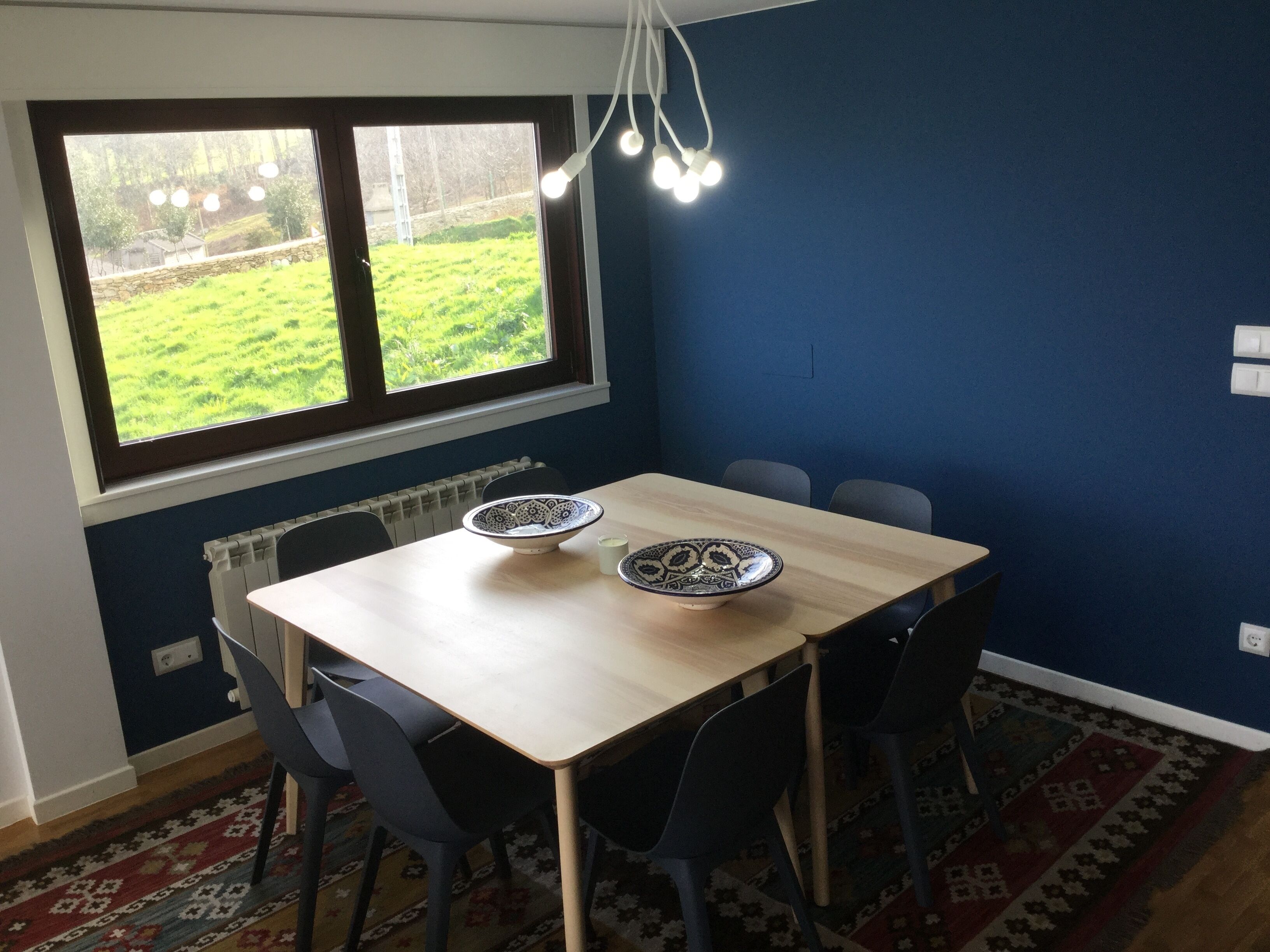 Ein Essbereich mit Tisch, Stühlen, einem Fenster mit Blick auf Grünflächen, einem Heizkörper und einem gemusterten Teppich.