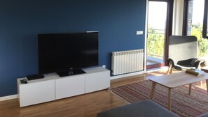 Ein modernes Wohnzimmer mit einem Flachbildfernseher auf einem weißen Schrank, einem Heizkörper, einem Sessel, einem kleinen Tisch und Blick ins Grüne durch das Fenster.