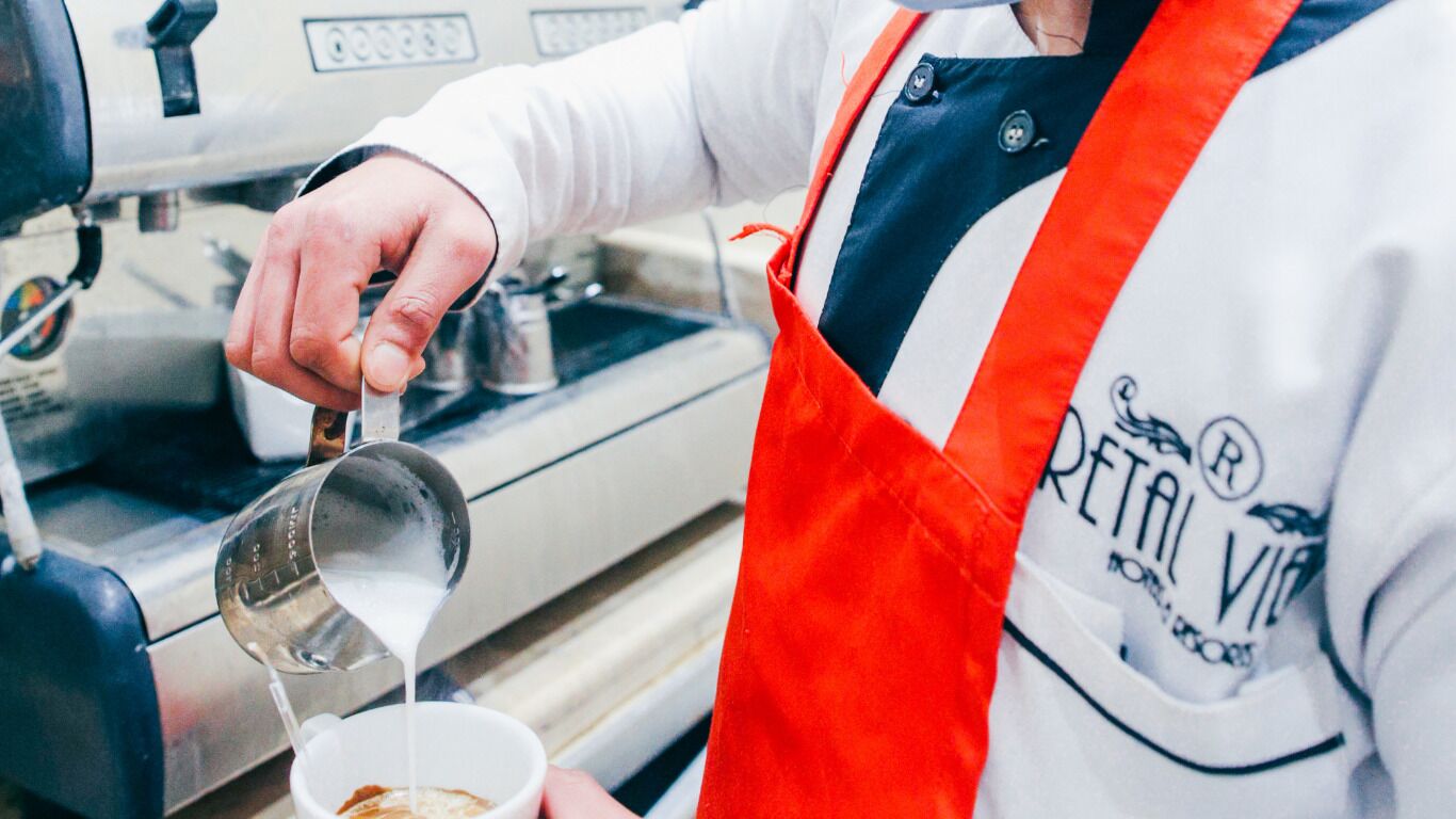 A person in a white shirt and red apron pouring milk into a cup.