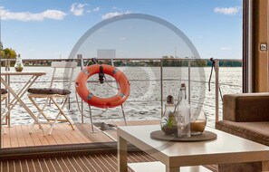 Une terrasse avec un gilet de sauvetage, une bouteille et un verre posés sur une table, donnant sur une étendue d’eau.