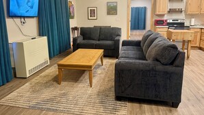 Una sala de estar con televisión, sofá, mesa de centro y una cocina con armarios y electrodomésticos.