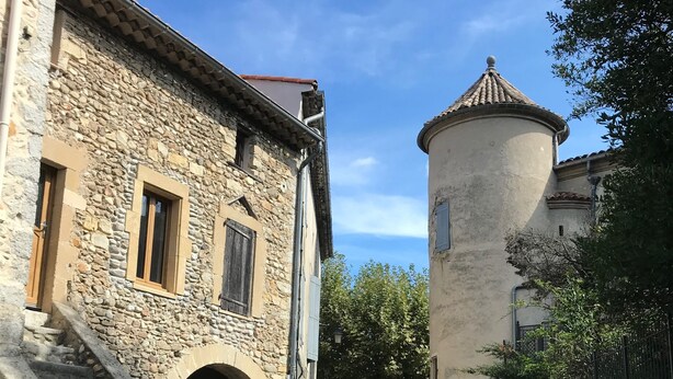 A narrow street flanked by stone buildings, one with a round tower and another with a staircase leading to an arched entrance.