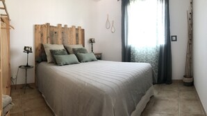 A bedroom with a wooden headboard, a bed with striped bedding, two bedside lamps, a window with curtains, and a tall plant beside the window.