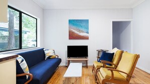 Una sala de estar con un sofá azul, sillones amarillos, una mesa de centro blanca y un televisor de pantalla plana sobre un soporte de madera.