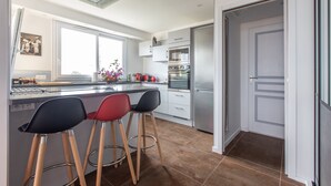 A modern kitchen with a central island, bar stools, stainless steel appliances, and a large window providing natural light. - Apartment panoramic view Baie des Anges (Nice)