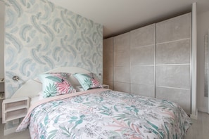 A bedroom with a floral-patterned bedspread, a nightstand, a lamp, and a wardrobe with a light-colored tiled wall. - Apartment panoramic view Baie des Anges (Nice)
