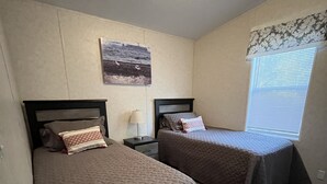 A room with two single beds, a small table with a lamp, a wall-mounted picture, and a window with blinds.