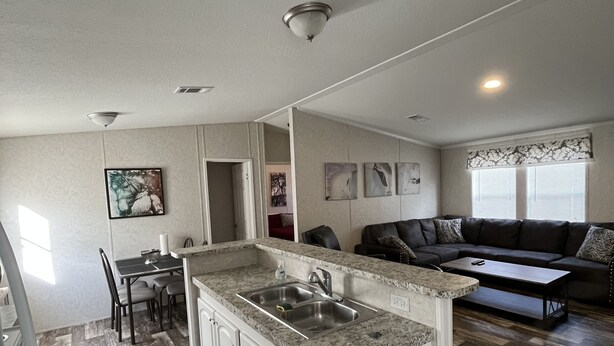 A modern kitchen with granite countertops, white cabinetry, and a central island with a sink. There is a dining area with a table and chairs, and a living area with a sofa and a coffee table.