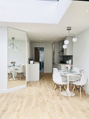 A modern kitchen with white cabinetry, a central island, and a dining area with white chairs and a round table. - AIR CON Modern 2BR House w/ Garden Near Beach (Whitstable)