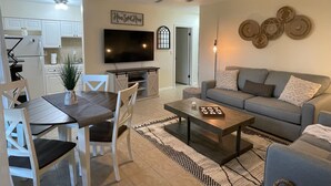 Ein modernes Wohnzimmer mit grauem Sofa, Couchtisch und Essbereich mit Tisch und Stühlen. Ein Flachbildfernseher ist an der Wand befestigt, darüber befindet sich ein Deckenventilator.