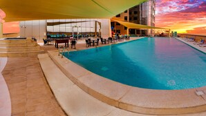 Piscina con sillas y mesas de descanso, rodeada por un edificio con vista a la puesta de sol.