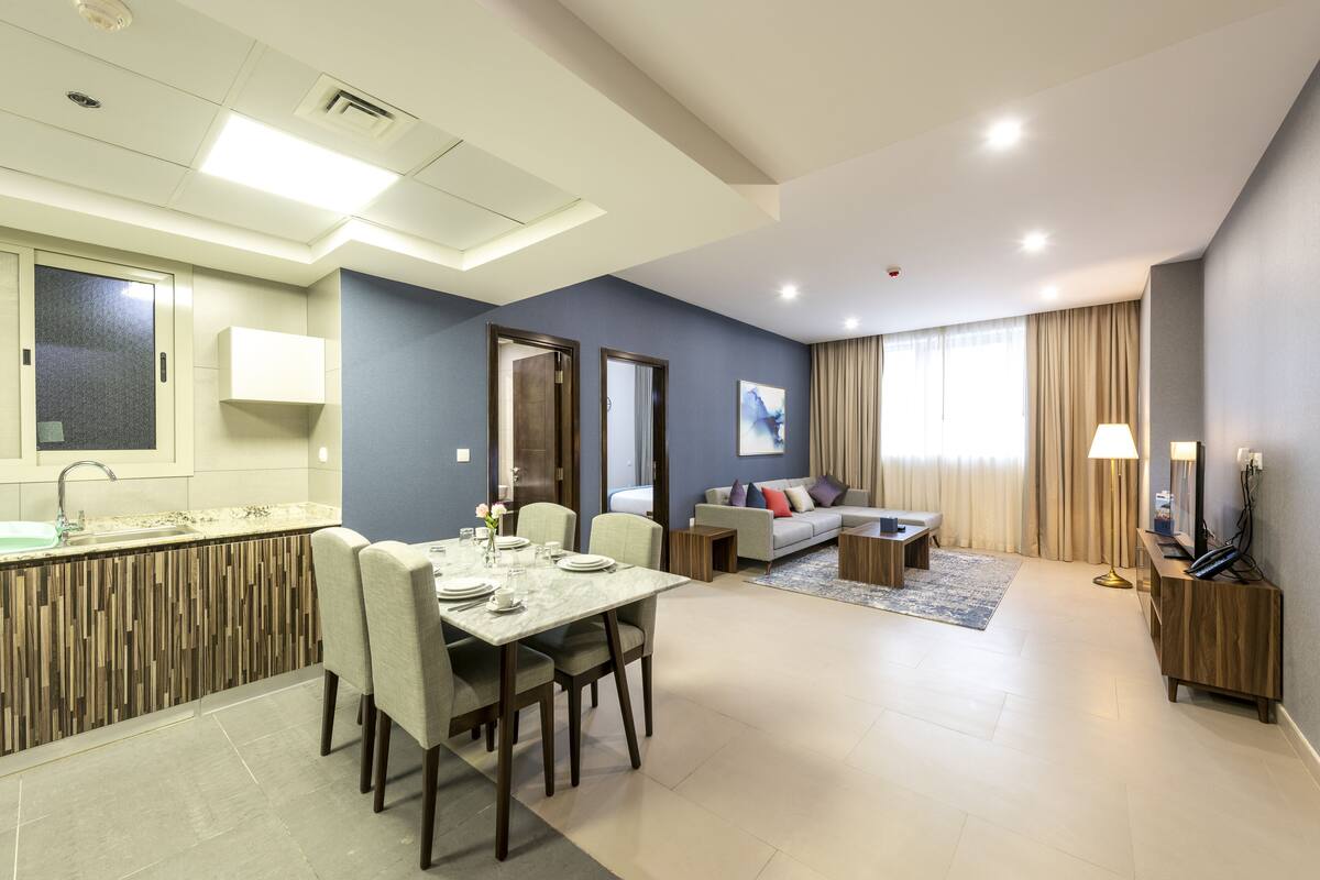 A modern hotel room with a dining area, a sofa, a TV, and a bathroom.