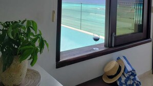 Una habitación con un ventanal grande que da a una piscina y al océano. Hay una planta en maceta sobre una mesa blanca y un sombrero de paja en un banco.