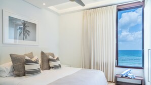 Un dormitorio con una cama grande, un cuadro enmarcado de una palmera, una ventana con vistas al océano y un banco con toallas.