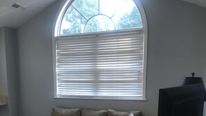 Ein Wohnzimmer mit einem Ecksofa, einem Fernseher und einem großen Fenster mit Jalousien.