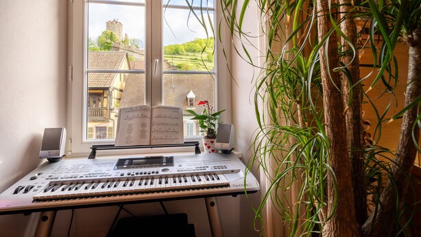 Un clavier avec des partitions, un haut-parleur et une plante près d’une fenêtre donnant sur des immeubles et une tour.