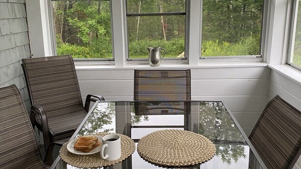 Un solarium con una mesa de vidrio, sillas de mimbre y vistas a un bosque.