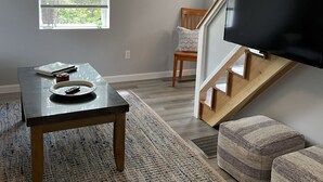 Una sala de estar moderna con una mesa de madera, un televisor montado en la pared, una escalera y vistas al exterior a través de una ventana con persianas.