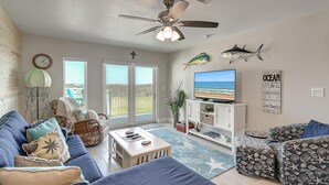 A living room with a large TV, a ceiling fan, and a view of the ocean through the sliding doors. - Poolside condo with covered patio, and direct pedestrian boardwalk to the beach! (Port Aransas)