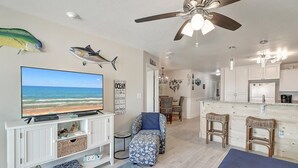 A living room with a TV displaying a beach scene, a ceiling fan, and a kitchen area with bar stools. - Poolside condo with covered patio, and direct pedestrian boardwalk to the beach! (Port Aransas)