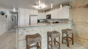 A kitchen with white cabinets, a granite countertop, a built-in island with bar stools, and pendant lighting. - Poolside condo with covered patio, and direct pedestrian boardwalk to the beach! (Port Aransas)