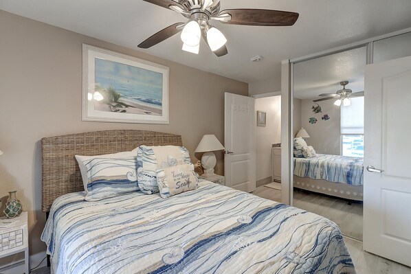 A bedroom with a bed, a ceiling fan, a painting, and a door leading to another room.