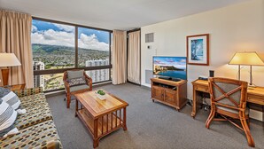 Ein Hotelzimmer mit großem Fenster und Blick auf die Stadt, einer Couch, einem Holztisch, einem Fernseher und einem Schreibtisch mit Lampe.