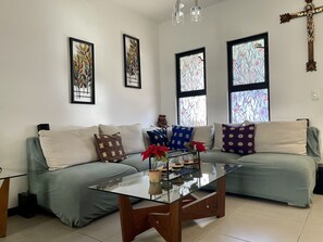 A living room with a sofa, a glass coffee table, and decorative pillows. There are two framed artworks on the wall and a wooden cross.