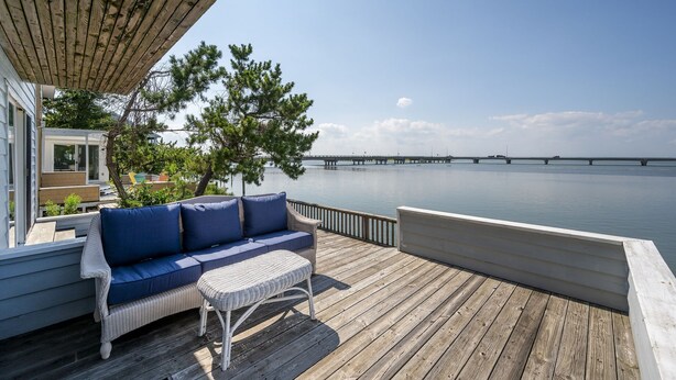 Una terraza de madera con un sofá de mimbre y una mesita, con vistas a un cuerpo de agua tranquilo y un puente.