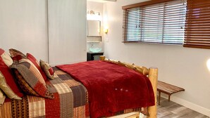Een slaapkamer met een houten bedframe, een rode deken en een sprei. Er staat een houten bankje, een raam met jaloezieën en een spiegel met verlichting erboven.