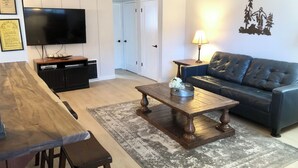 Een woonkamer met een houten salontafel, een zwarte leren bank, een televisie op een meubel, een lamp en een deur naar een andere kamer.