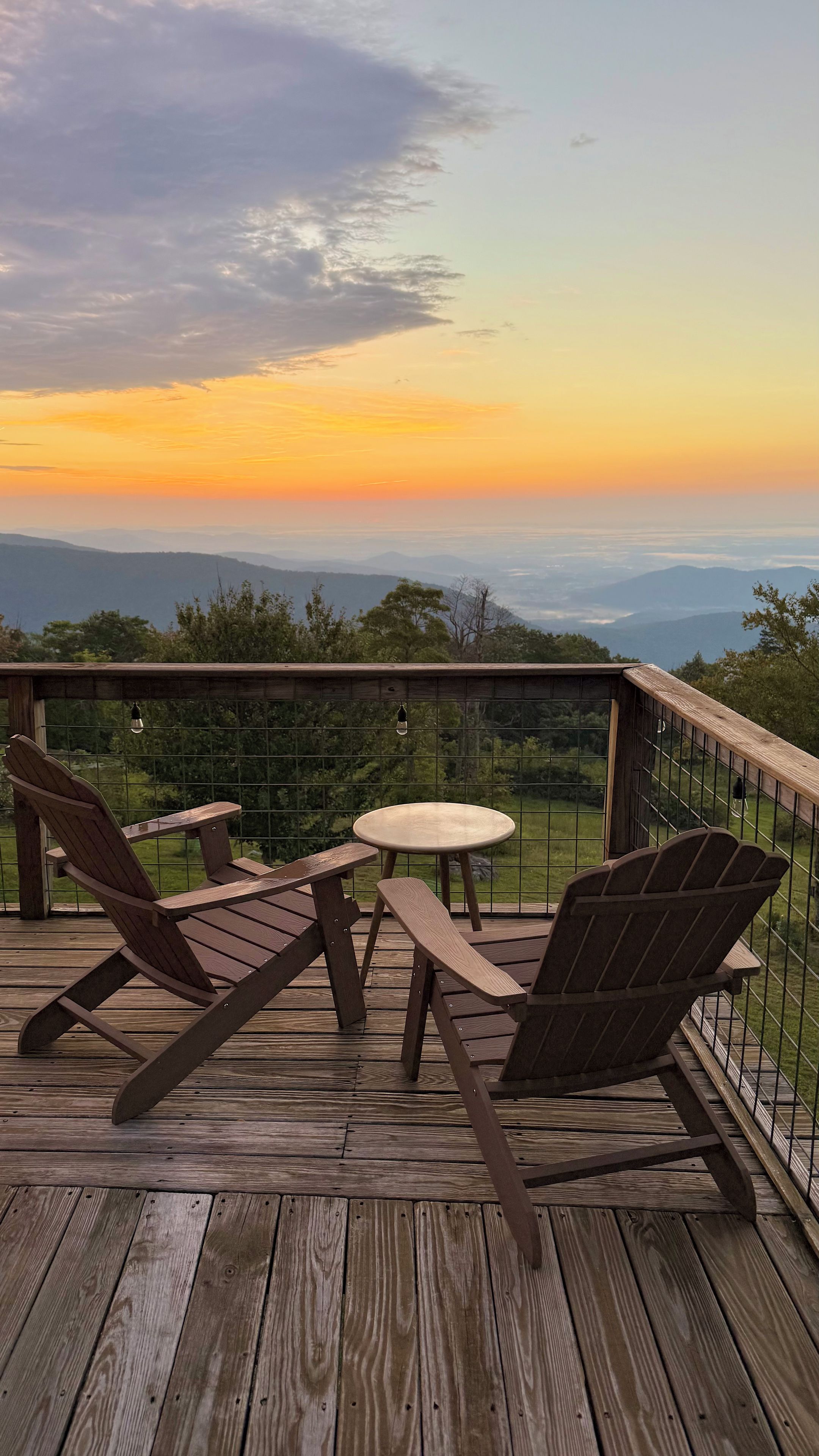 Una terraza de madera con sillas Adirondack y una mesa pequeña, con vista a un paisaje montañoso al atardecer.